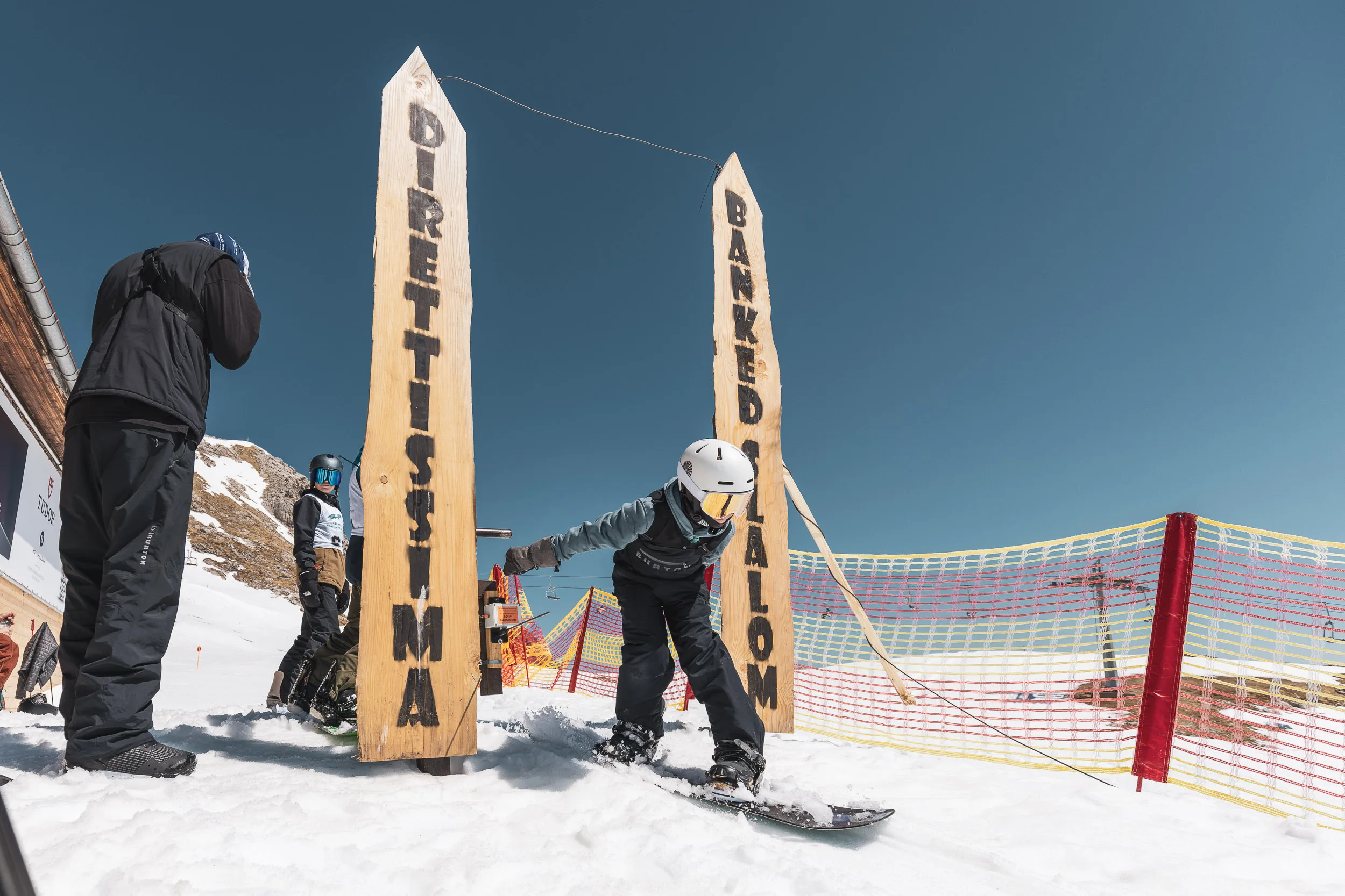 Zwei Rider am hölzernen Startbogen „Direttissima – Banked Slalom“ vor blauem Himmel am Nebelhorn