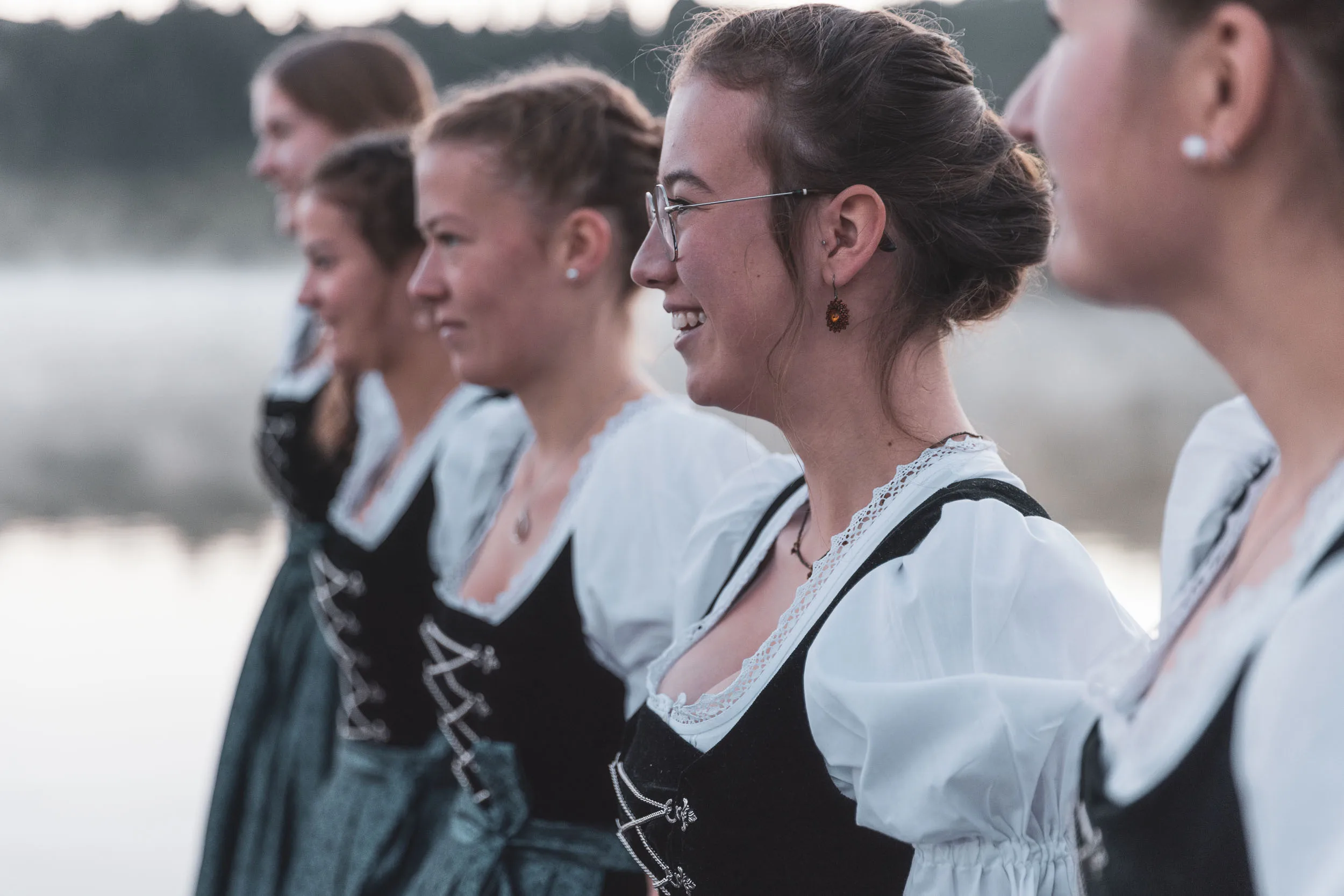Tänzerinnen in Tracht stehen am Alpsee vor einer ruhigen Nebelkulisse.