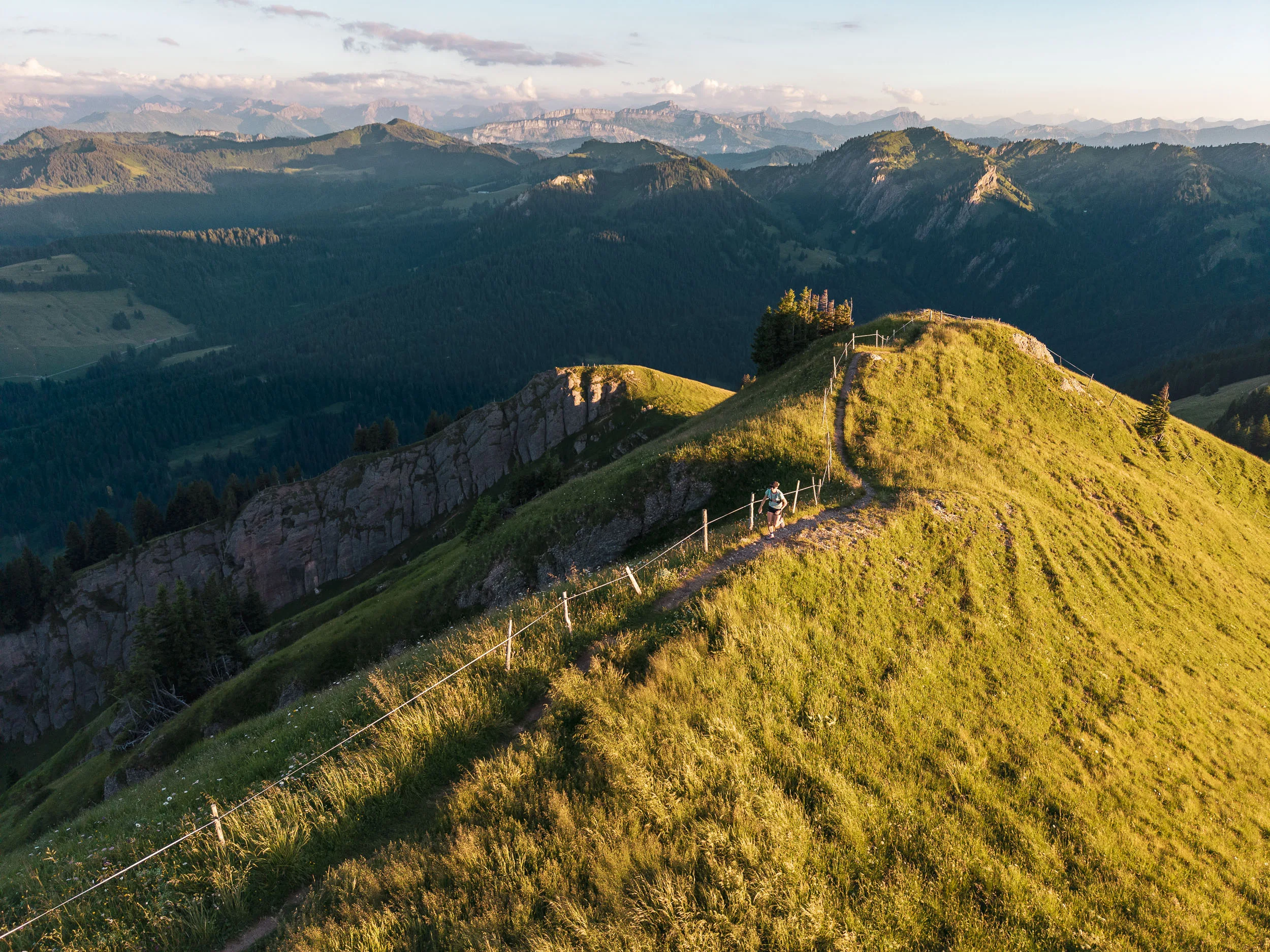 Eine Trailrunnerin folgt einem sonnigen Gratweg oberhalb schattiger Täler und bewaldeter Berghänge.