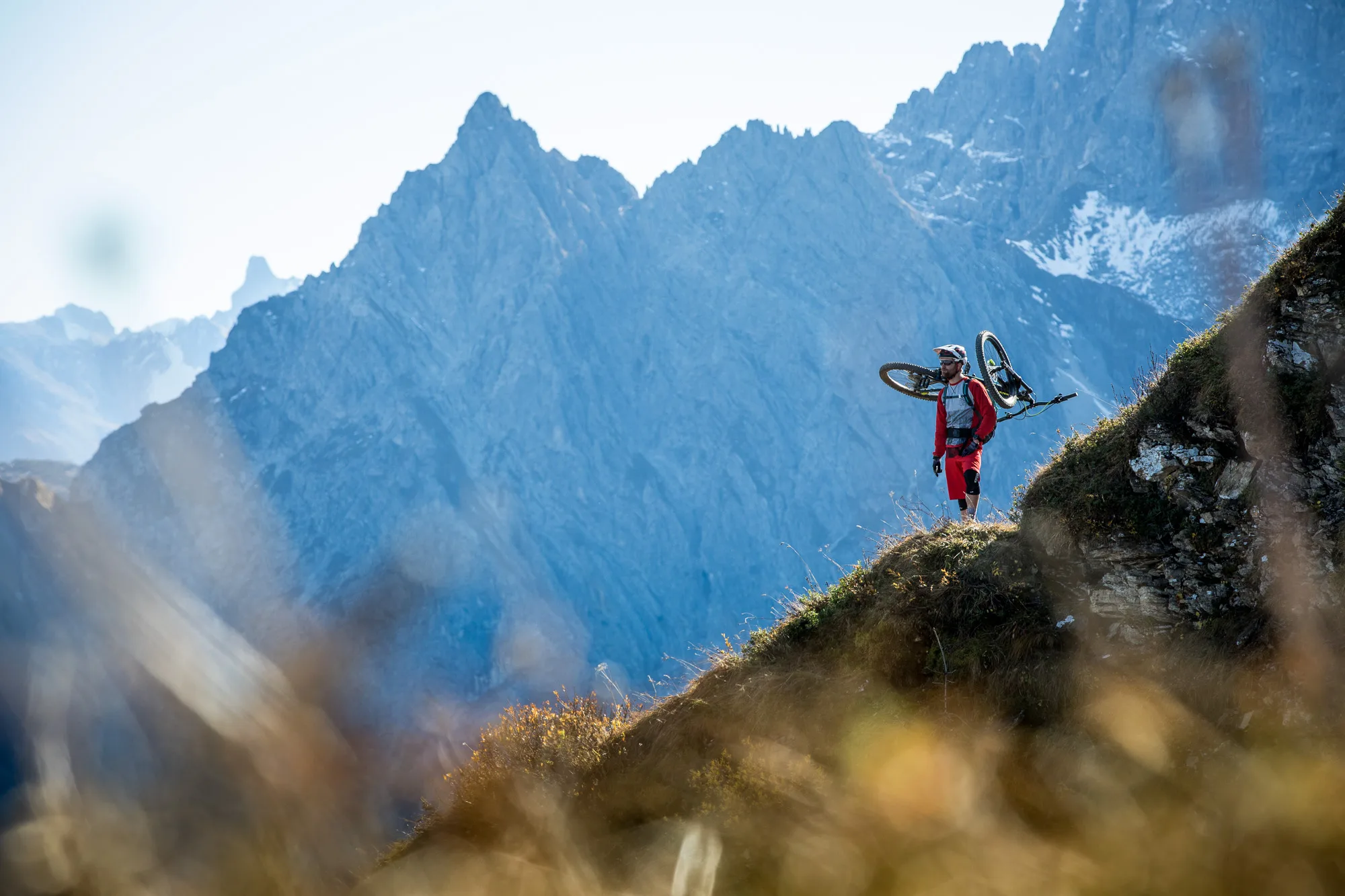 Mountainbiker mit geschultertem Rad auf einer felsigen Kante vor hohen Alpengipfeln