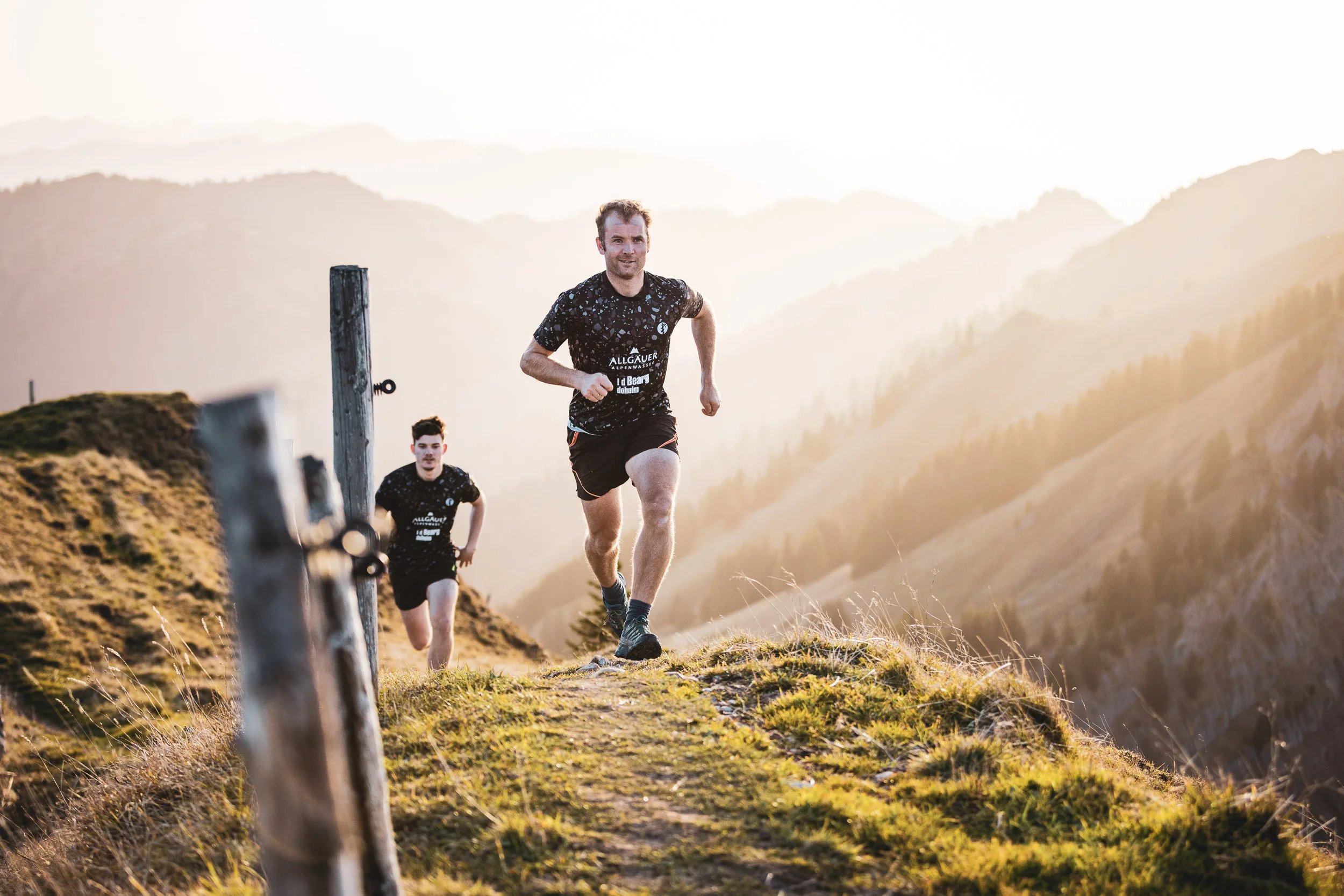 Trailrunner laufen dynamisch an Zaunpfosten entlang ueber einen Berggrat