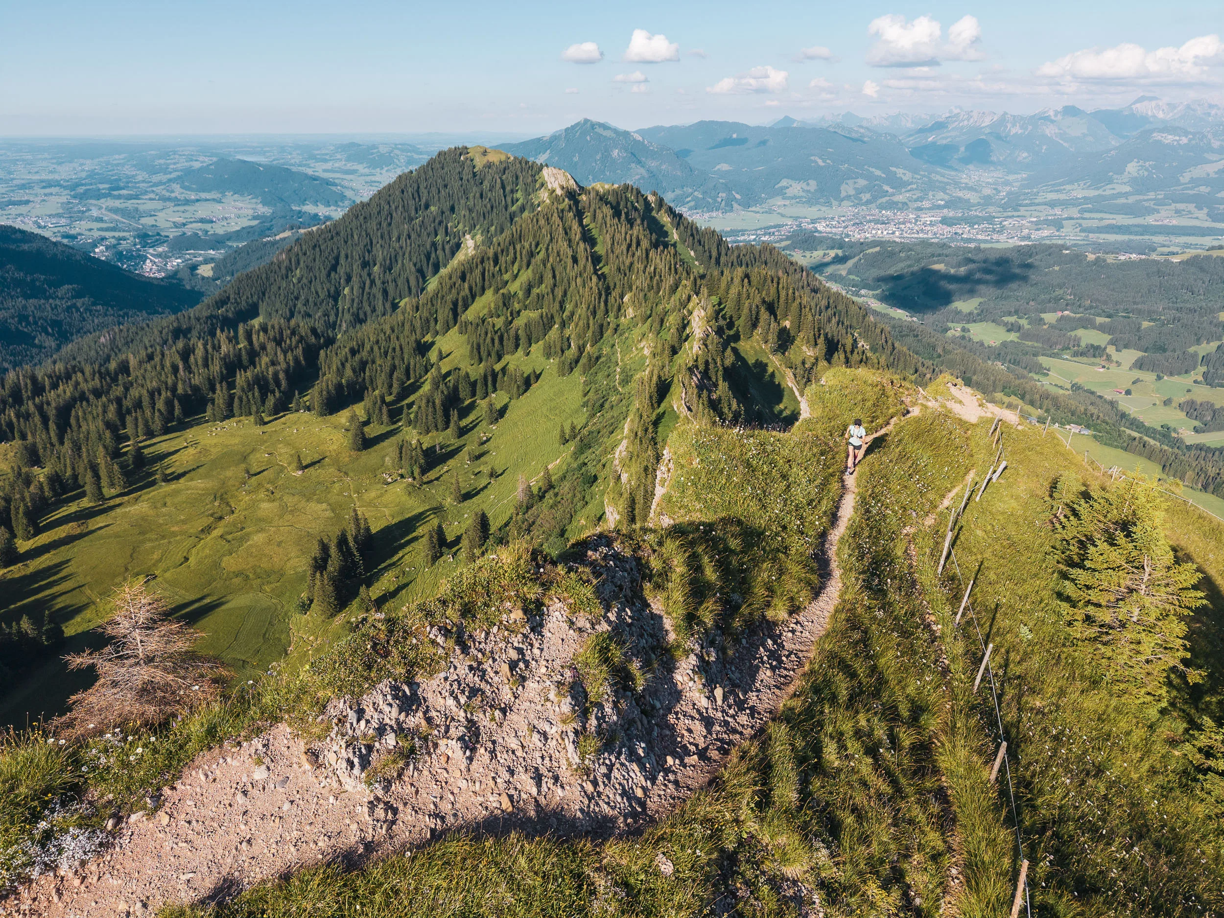 Eine Trailrunnerin folgt einem schmalen Höhenweg mit Blick auf bewaldete Berge und das weite Tal.