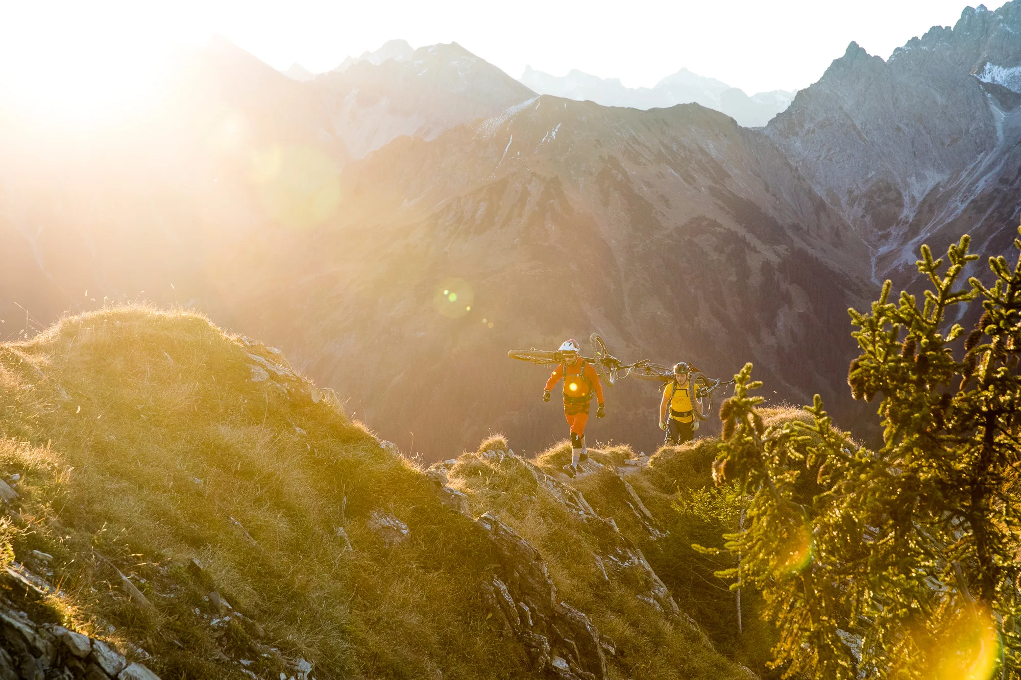 Drei Mountainbiker tragen ihre Räder über einen alpinen Grat im warmen Gegenlicht