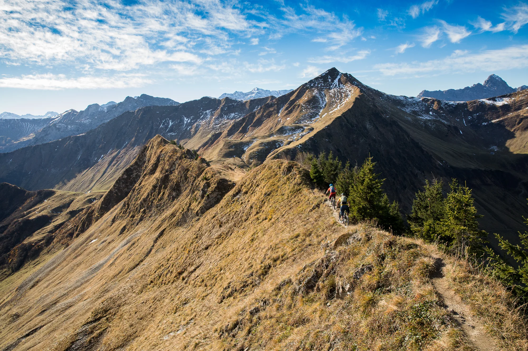 Zwei Mountainbiker fährt auf einem schmalen Gratweg durch die weite Alpenlandschaft
