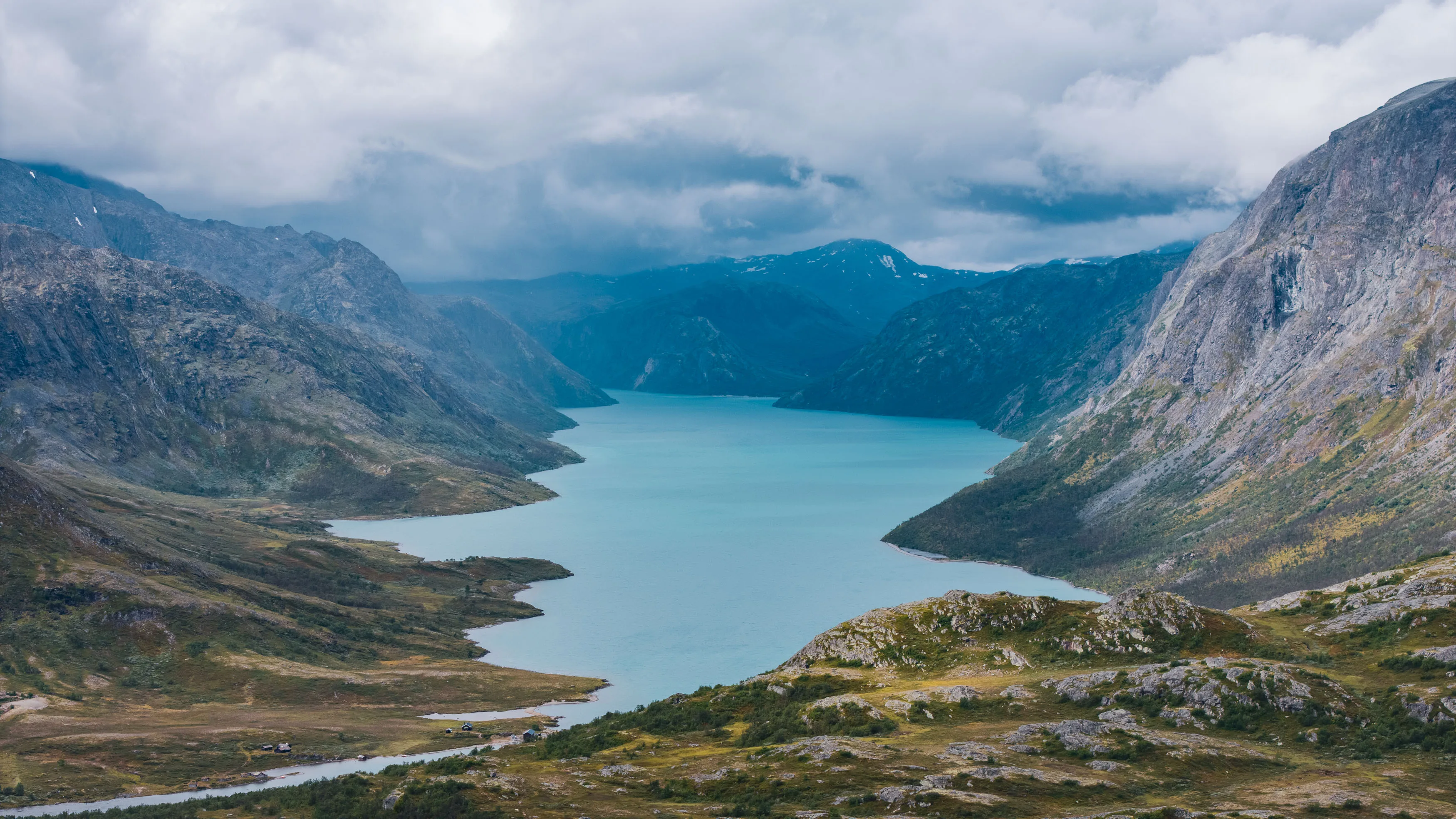 Türkisfarbener Bergsee liegt zwischen steilen felsigen Hängen unter tiefen Wolken