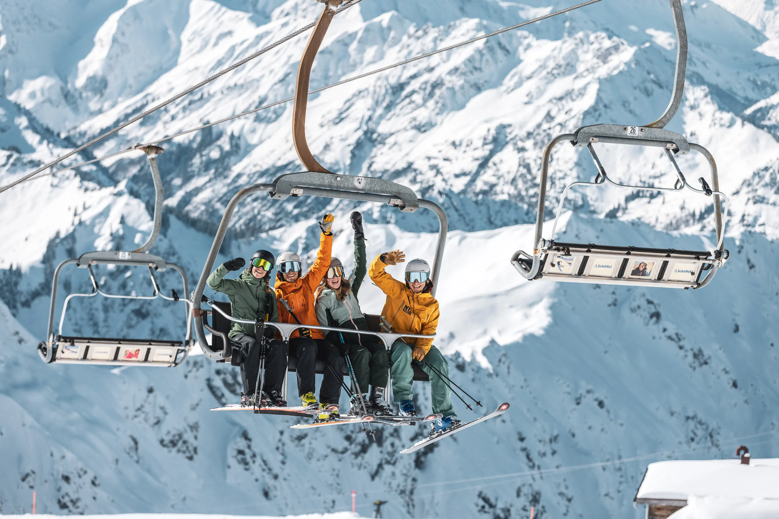 Drei Skilehrer winken aus einem Sessellift vor verschneitem Berghang