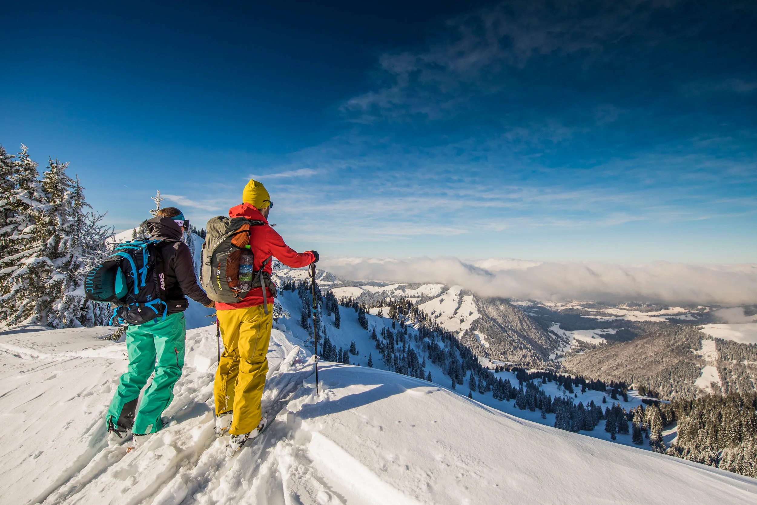 Zwei Skitourengeher stehen mit Ski auf einem sonnigen Aussichtspunkt.