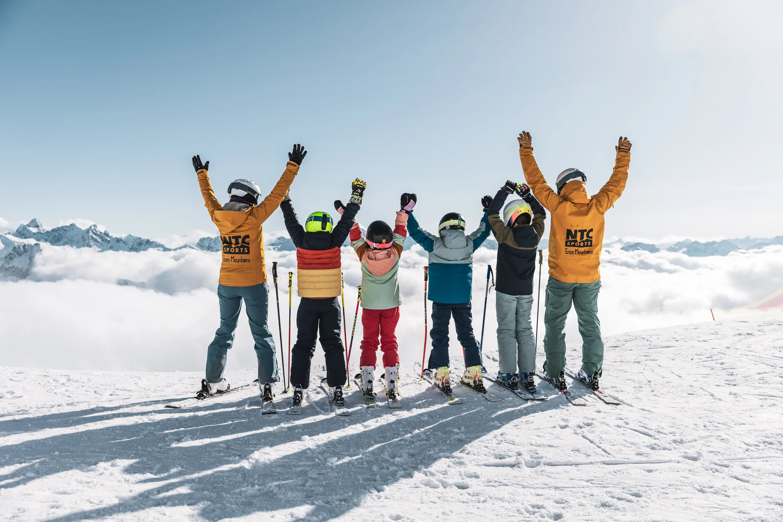 Skikurs und Lehrkräfte stehen mit erhobenen Armen vor dem Alpenpanorama