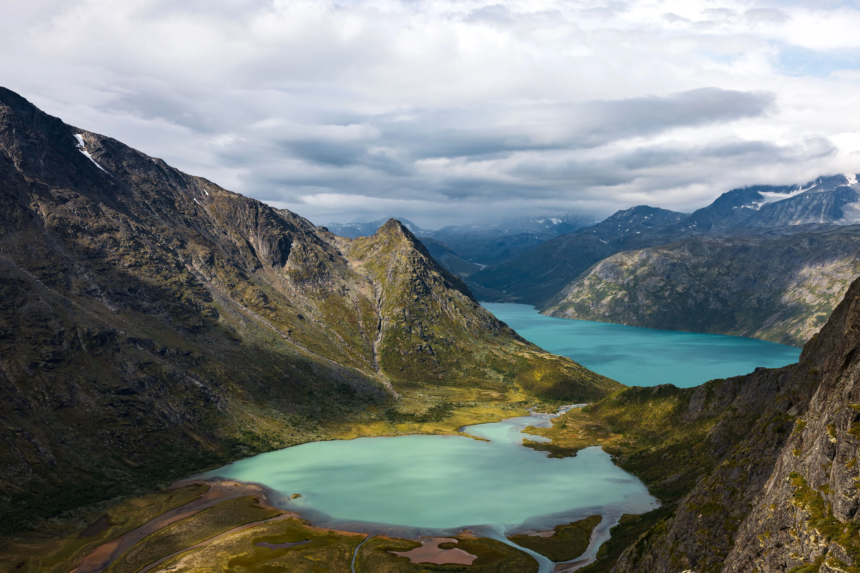 Heller Bergsee und verzweigter Fluss liegen zwischen schroffen Bergen unter grauen Wolken