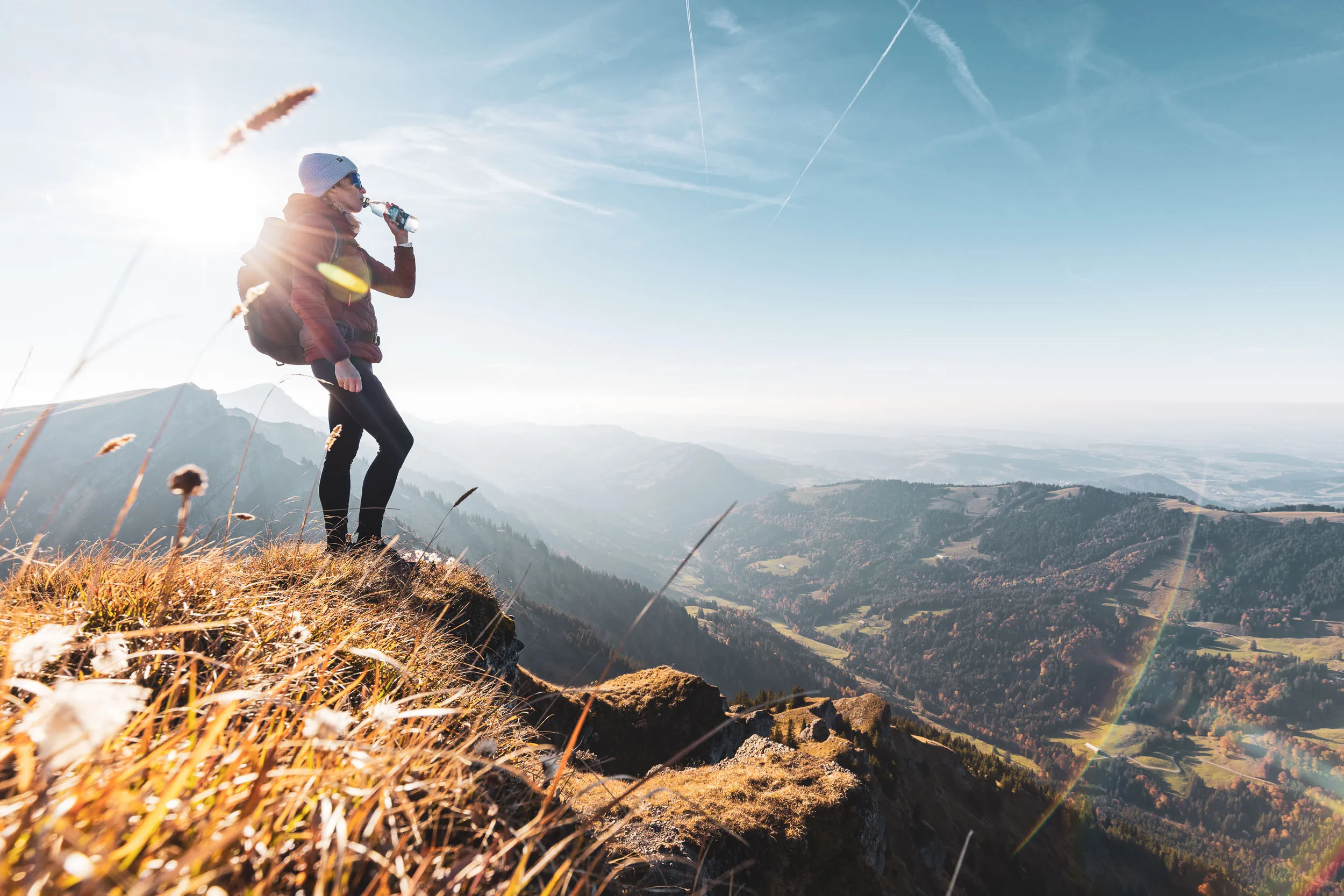 Wanderin trinkt im starken Gegenlicht auf einem grasigen Bergvorsprung