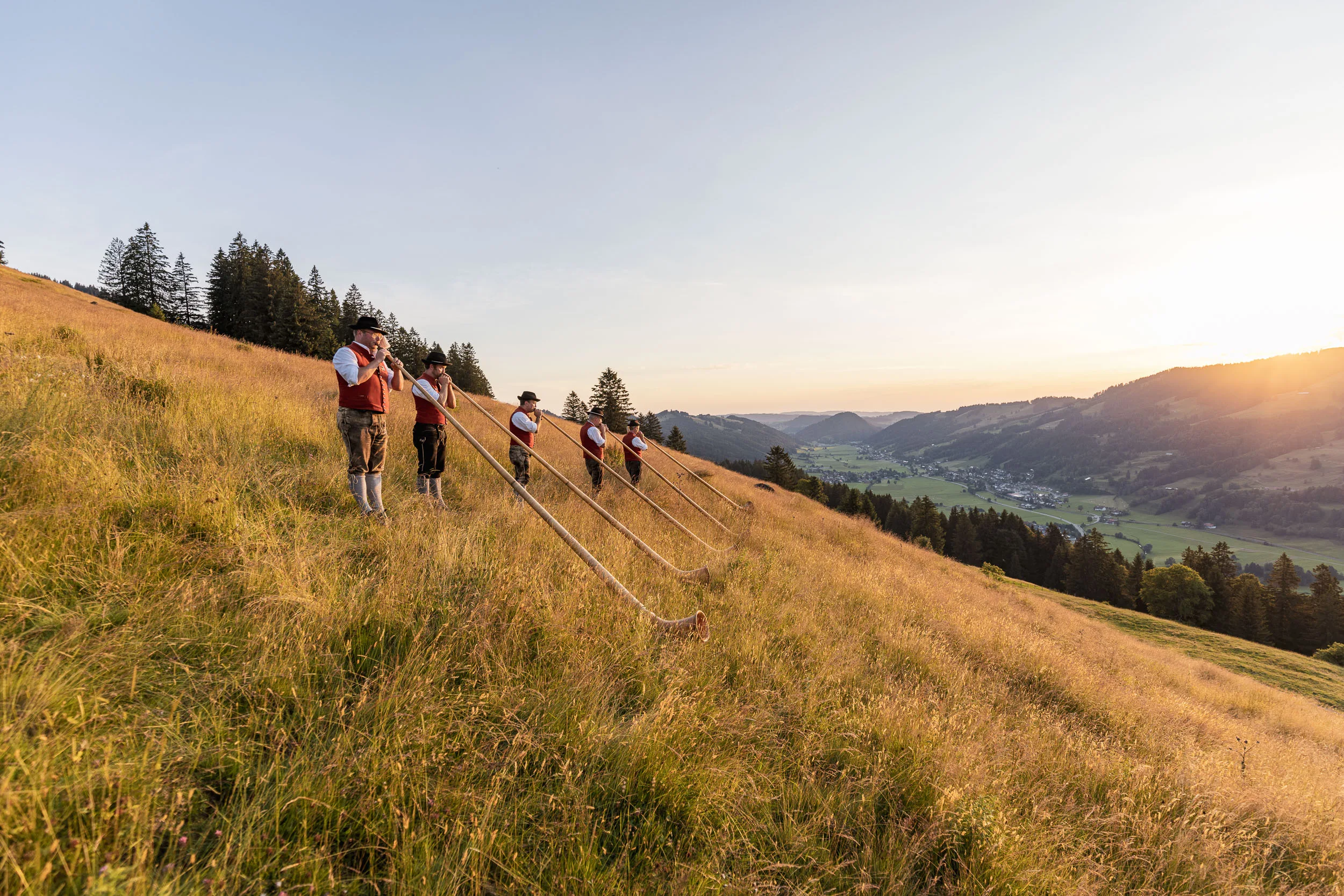 Alphornbläser auf einer weiten Bergwiese mit Abendsonne über dem Allgäu.