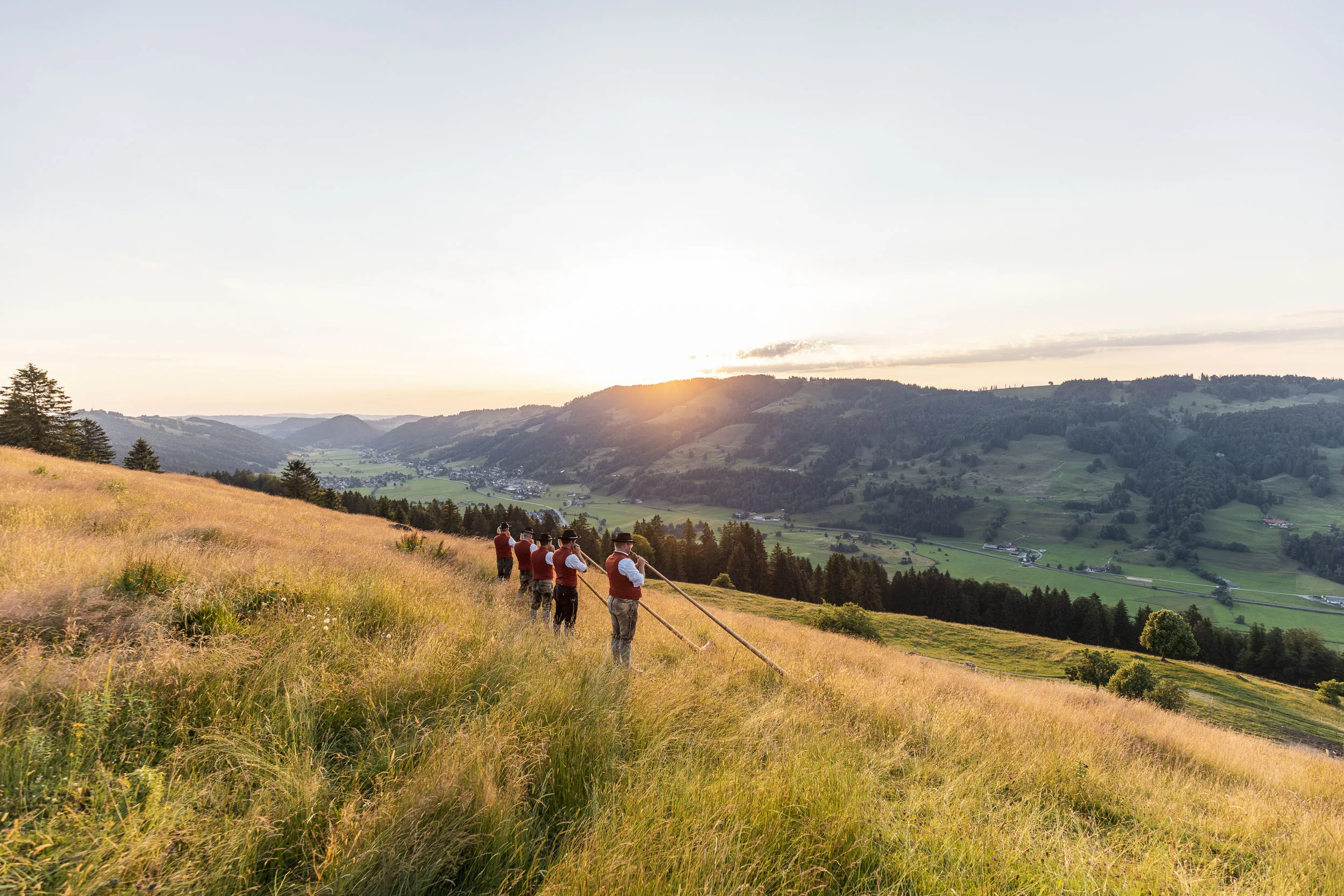 Alphornbläser auf einem sommerlichen Hang mit Blick in die Abendlandschaft.