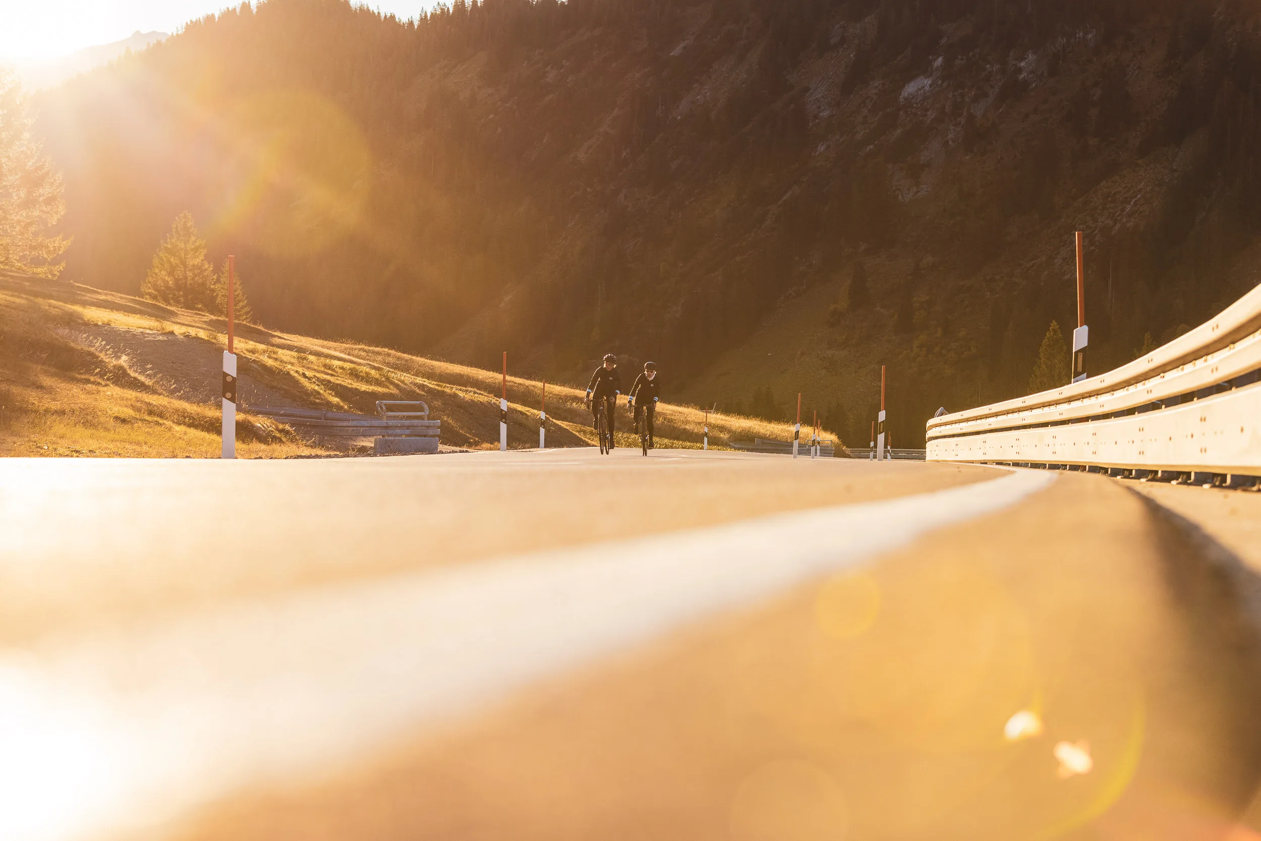 Zwei Rennradfahrerinnen auf einer weiten Passstraße im goldenen Licht