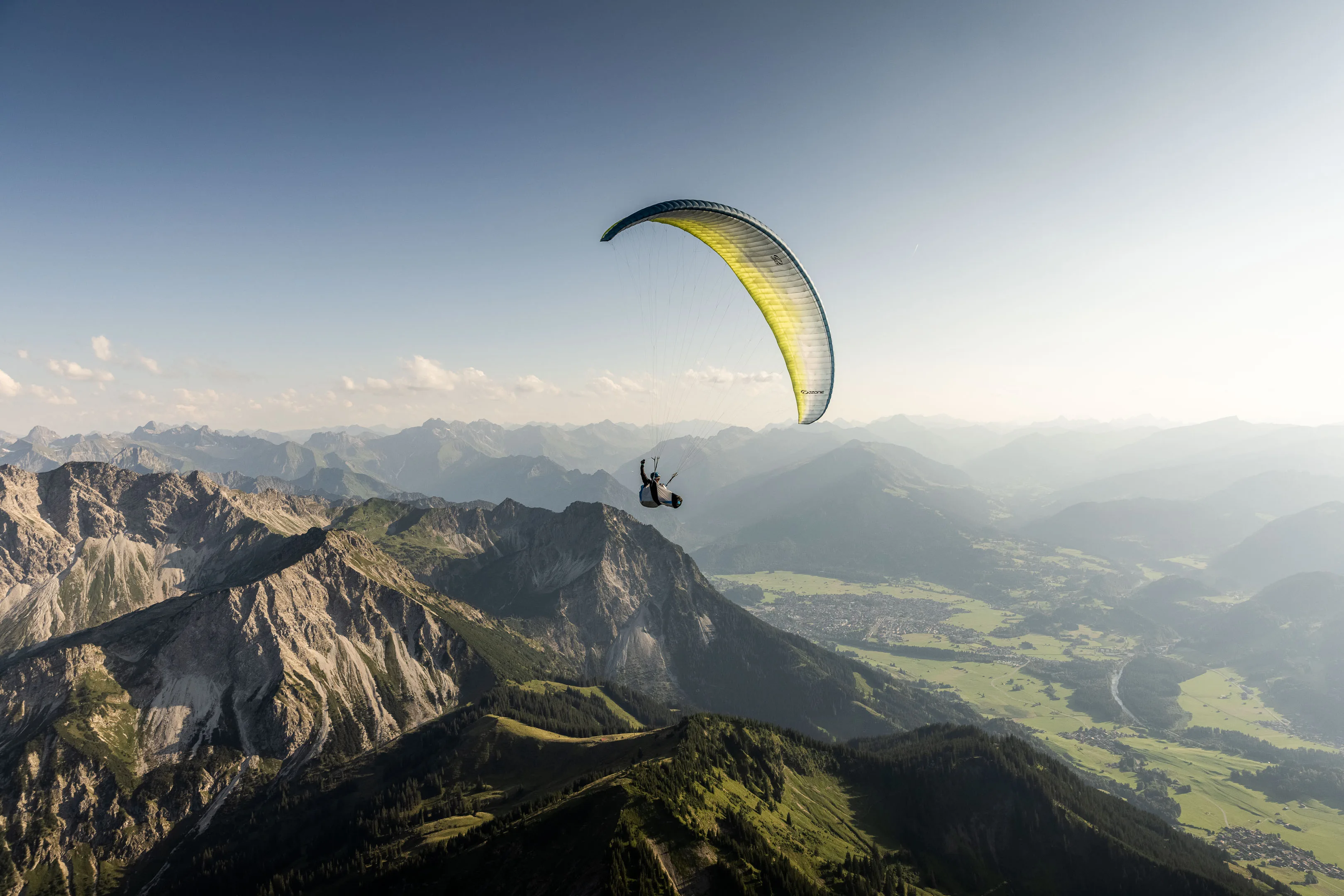 Ein Gleitschirm fliegt vor hellem Talgrund und gestaffelten Bergen in der Ferne