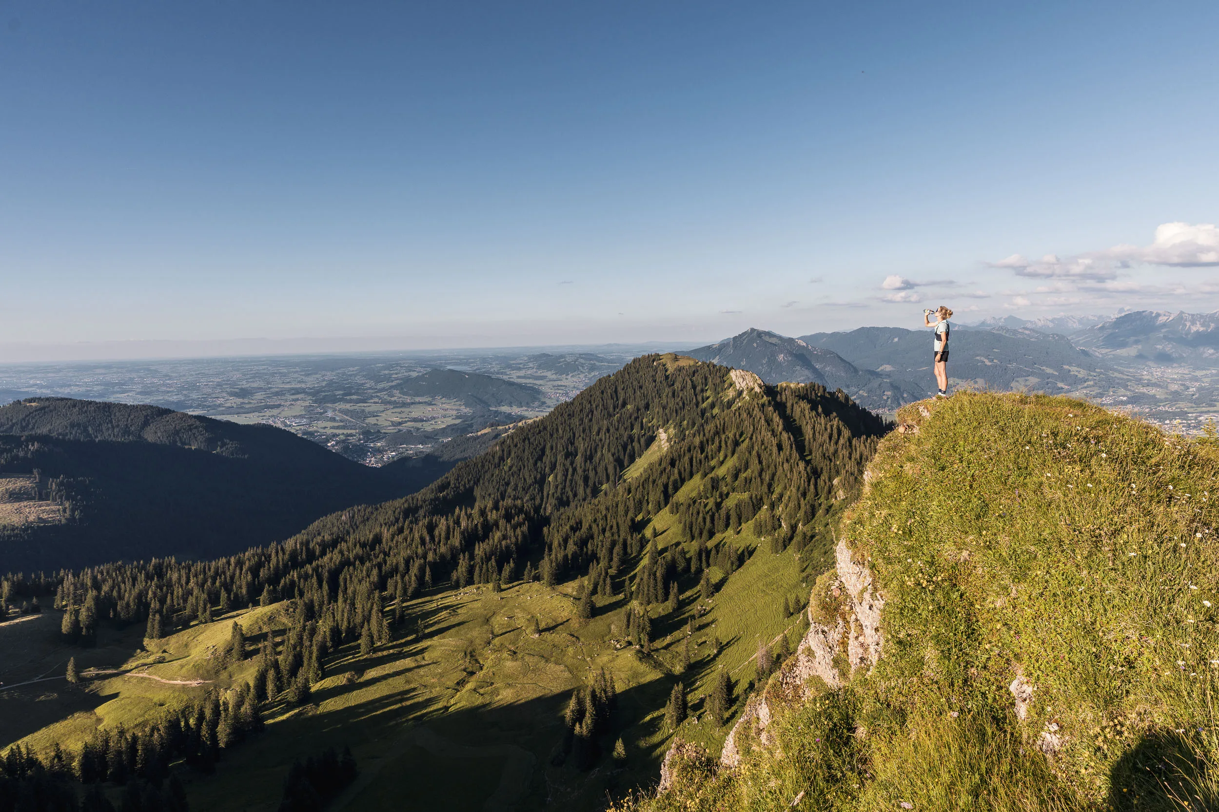 Eine Läuferin steht mit erhobener Wasserflasche auf einer Bergkuppe vor einem weiten Talpanorama.