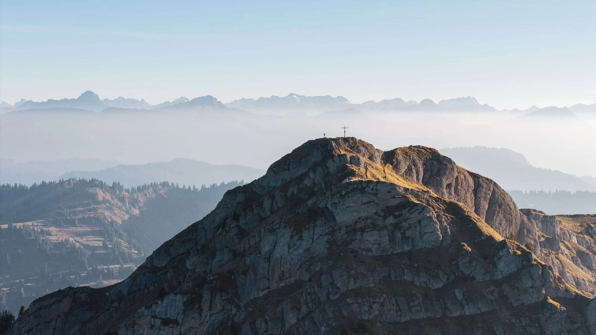 Ein Gipfel mit Kreuz steht über einem hellen Nebelband und fernen Bergsilhouetten