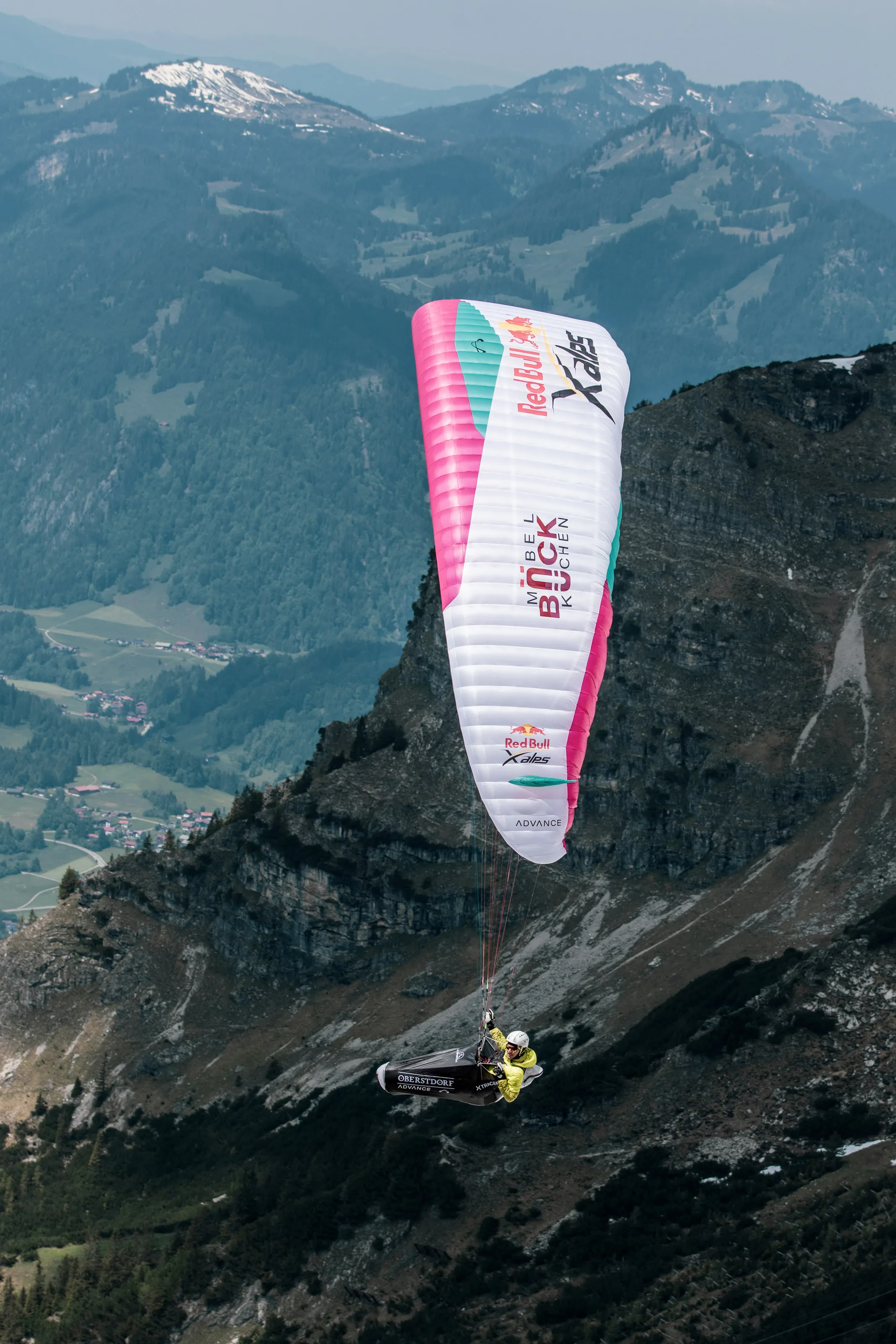 Im Hochformat fliegt Michael Lacher unter dem pinkweißen Schirm vor einer steilen Felsflanke.