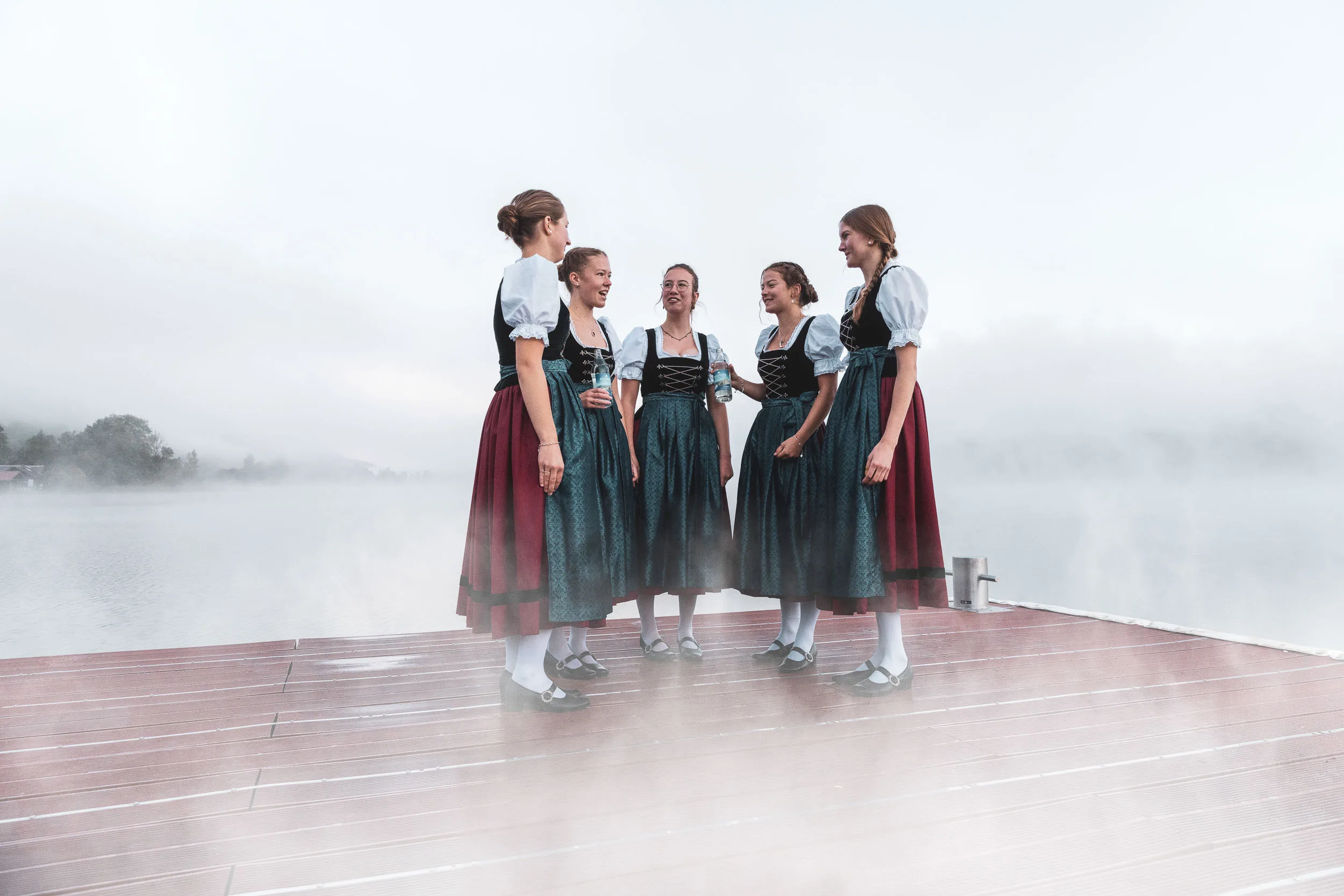 Tänzerinnen in Tracht auf einem Holzsteg am nebligen Alpsee.