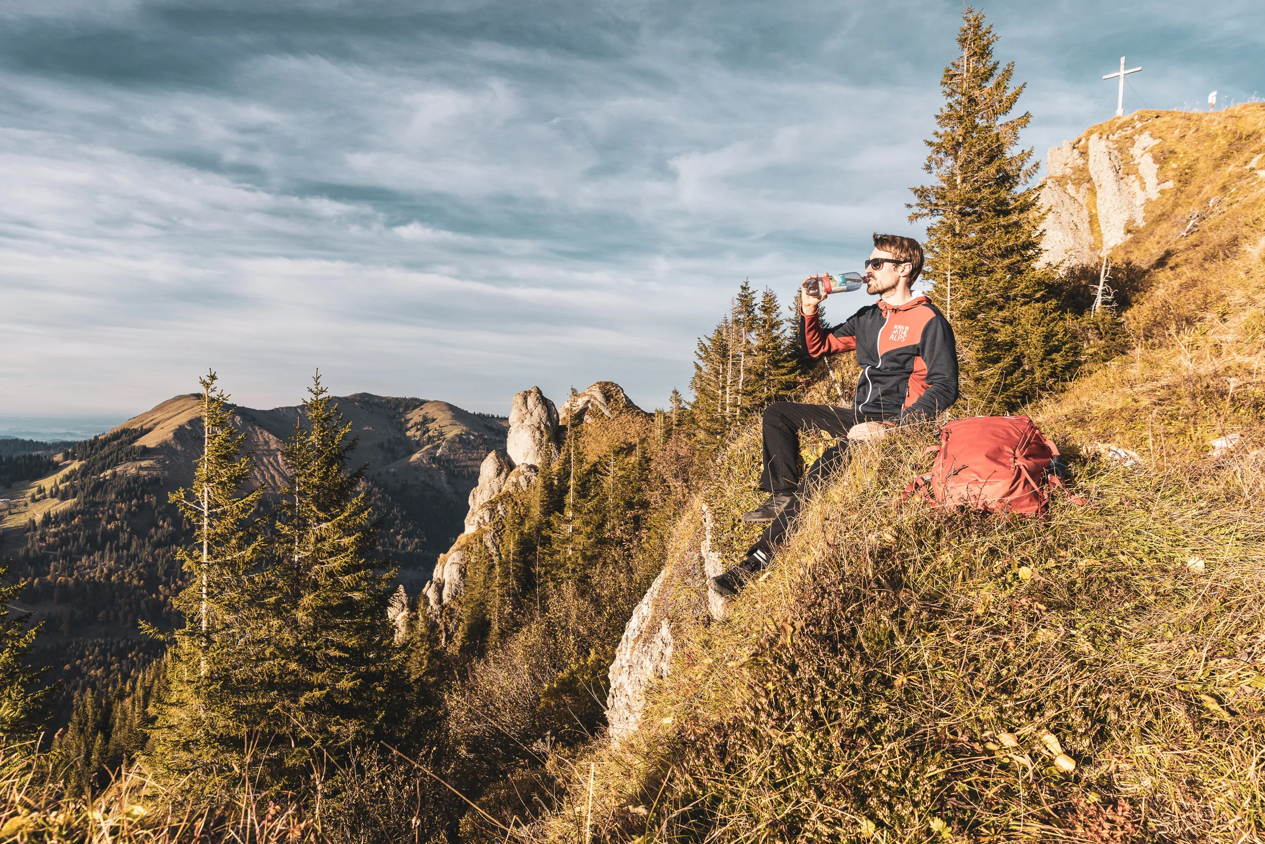 Andreas Kögel sitzt bei einer Trinkpause auf einer sonnigen Bergwiese über dem Tal