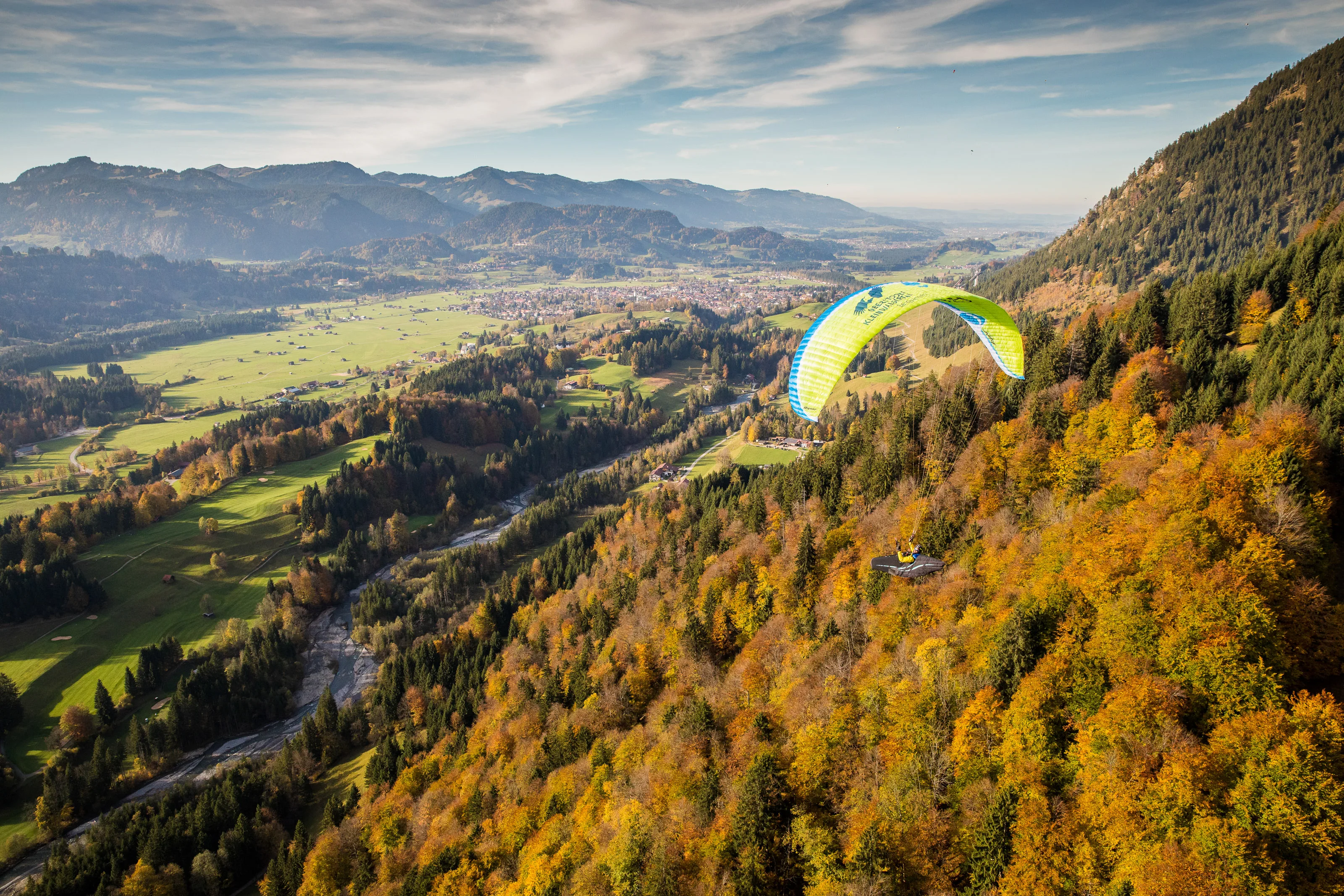 Michael Gebert fliegt mit grünem Gleitschirm am herbstlichen Waldhang mit Blick über das Oberstdorfer Tal