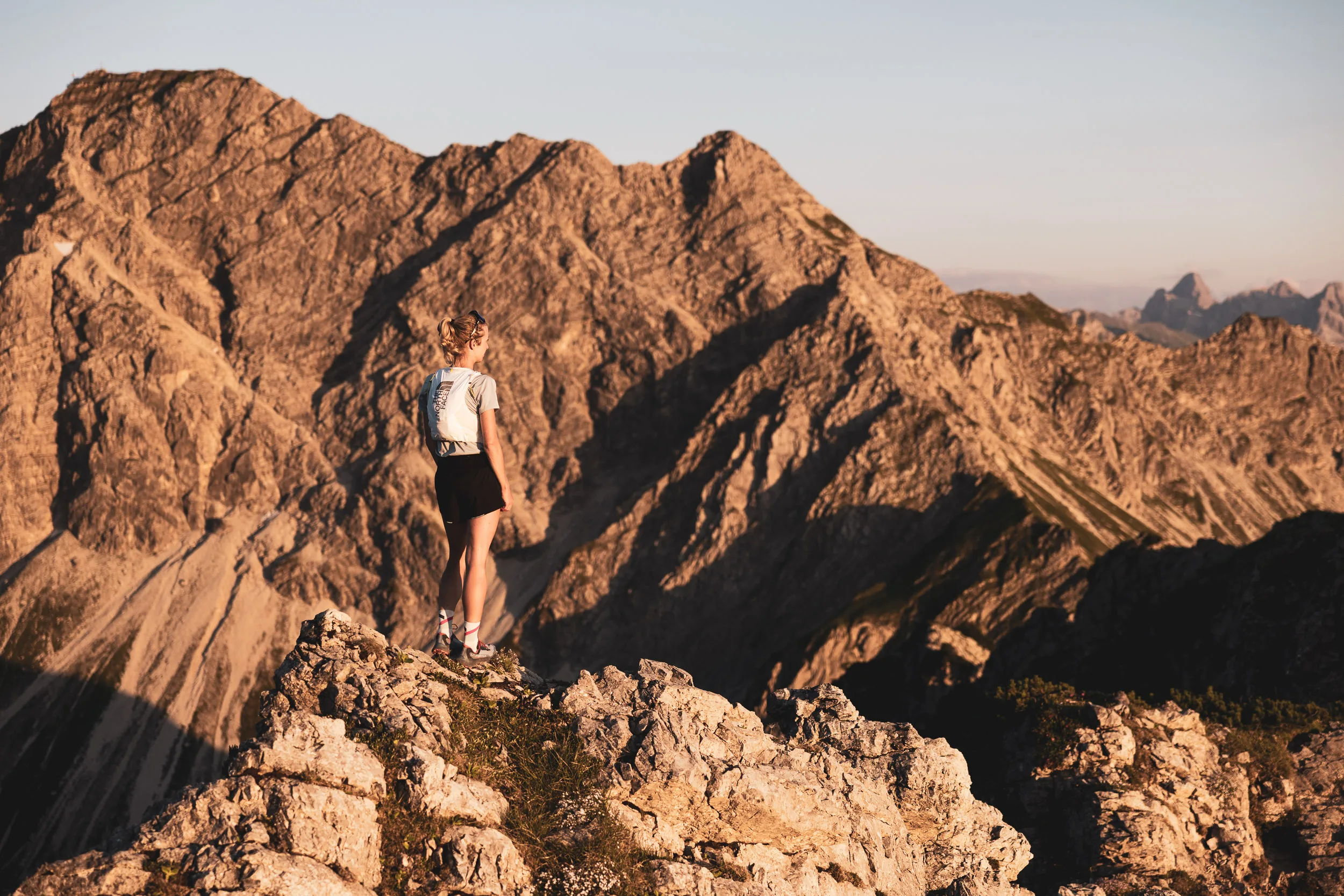 Trailrunnerin von hinten auf einem Felsgrat vor sonnenbeschienenen Bergen