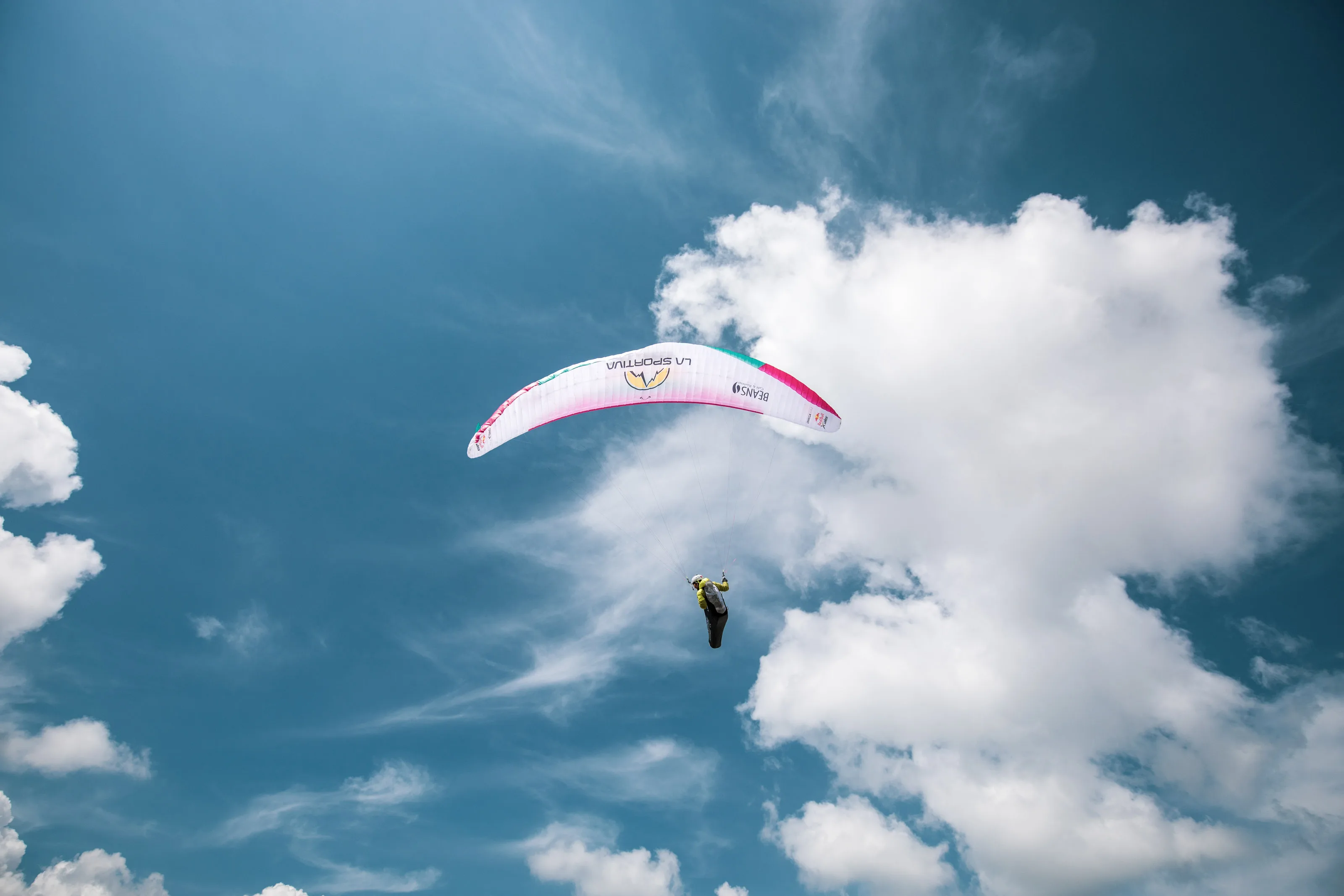 Der pinkweiße Gleitschirm mit Michael Lacher hebt sich vor blauem Himmel und hellen Wolken ab.