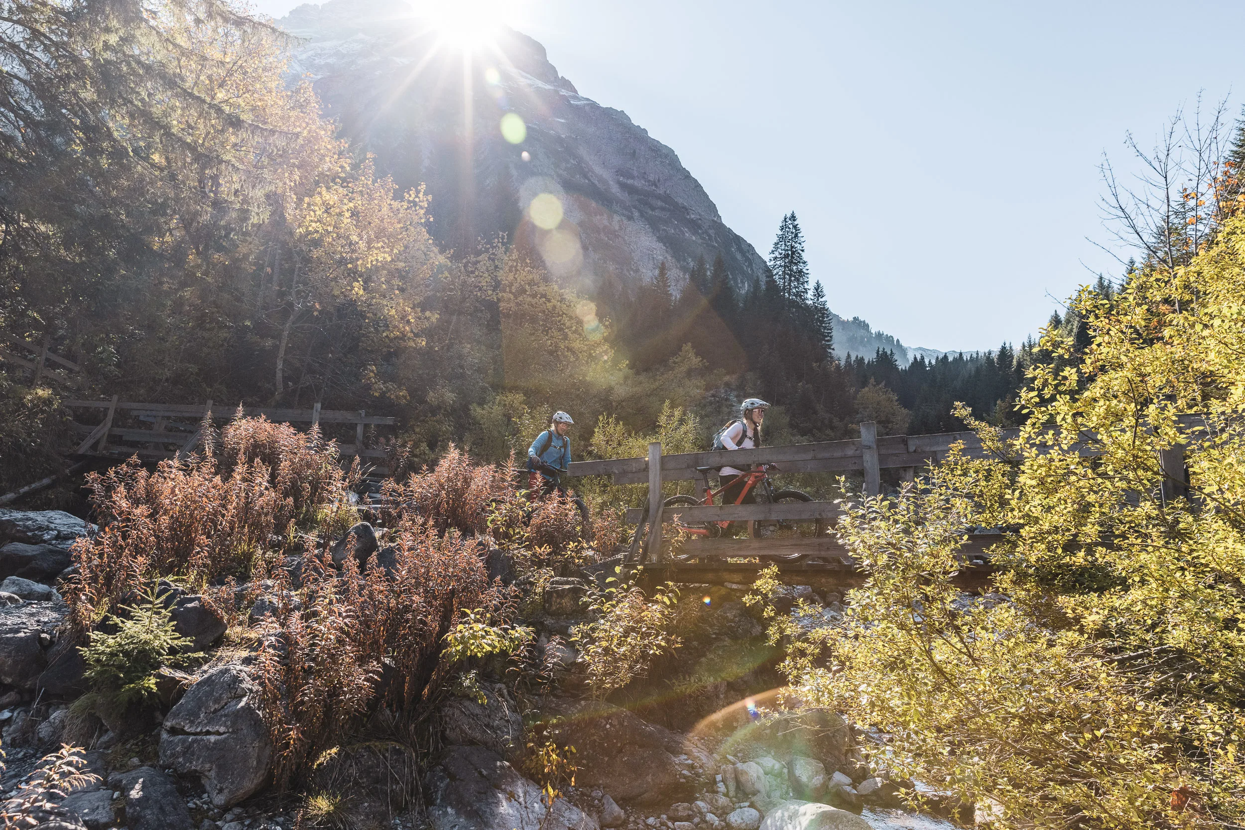 Silvi Vogel und Andrea Geiger schieben mit ihren Mountainbikes an einem Bach im Herbstlicht