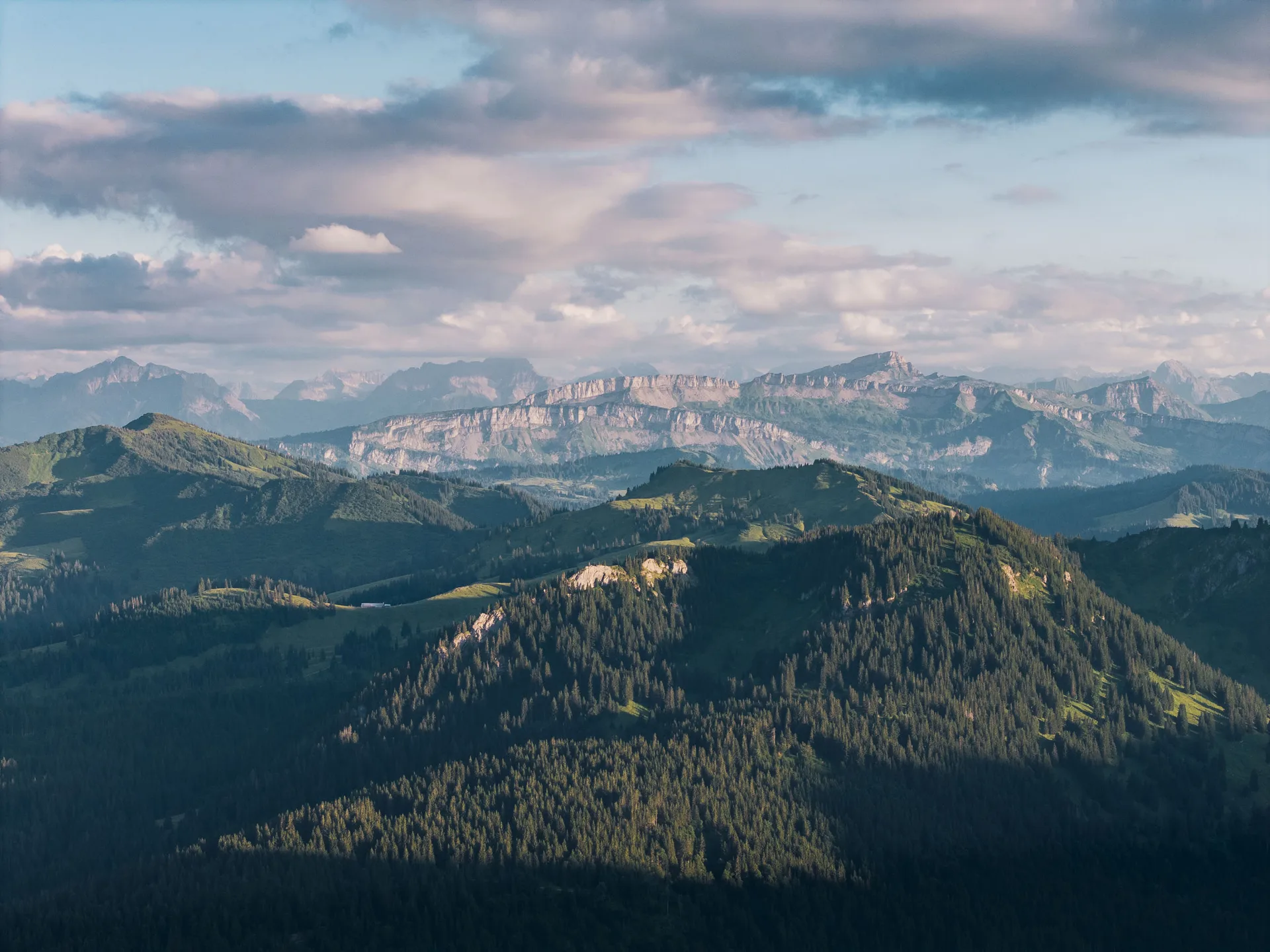 Bewaldete Bergrücken liegen vor hellen Felswänden und locker bewölktem Himmel im Abendlicht.