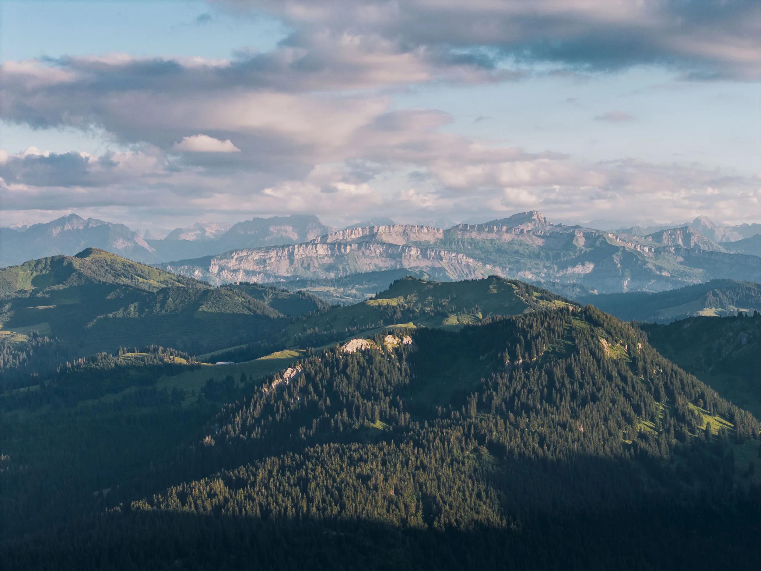 Bewaldete Bergrücken liegen vor hellen Felswänden und locker bewölktem Himmel im Abendlicht.