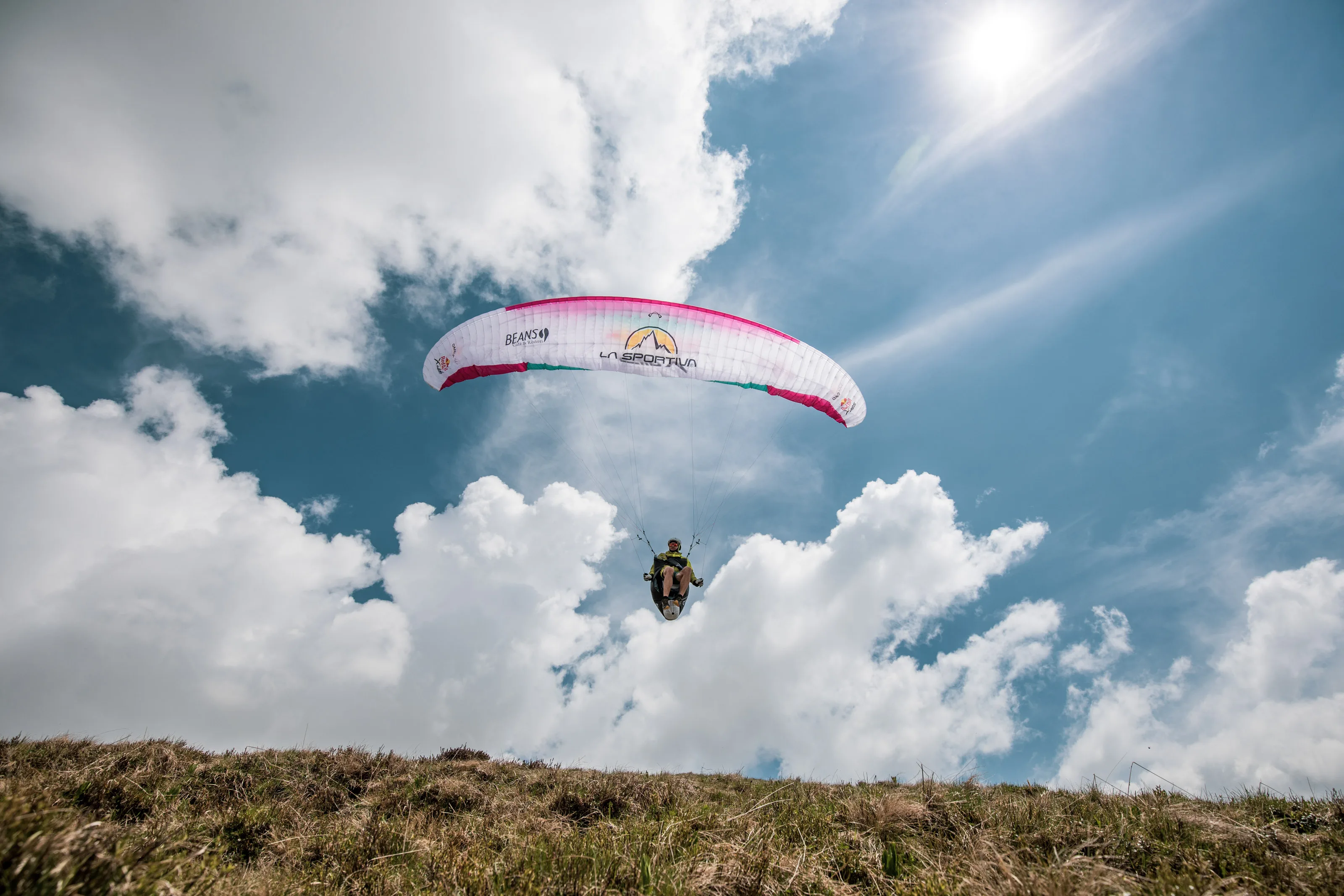 Michael Lacher fliegt unter dem pinkweißen Schirm vor großen weißen Wolken über einem Grashang.