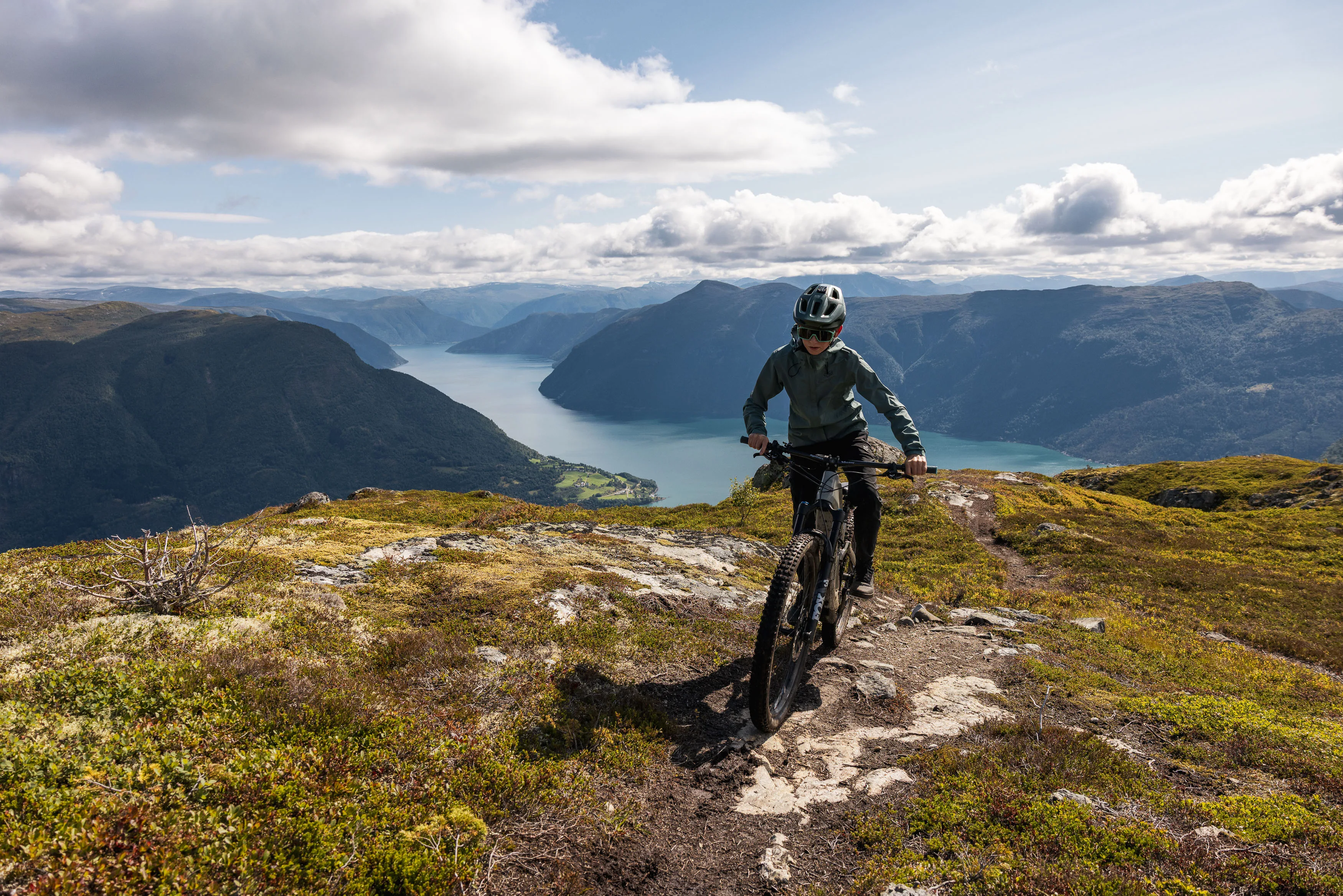 Mountainbiker fährt auf einem felsigen Trail mit weitem Fjordpanorama auf die Kamera zu