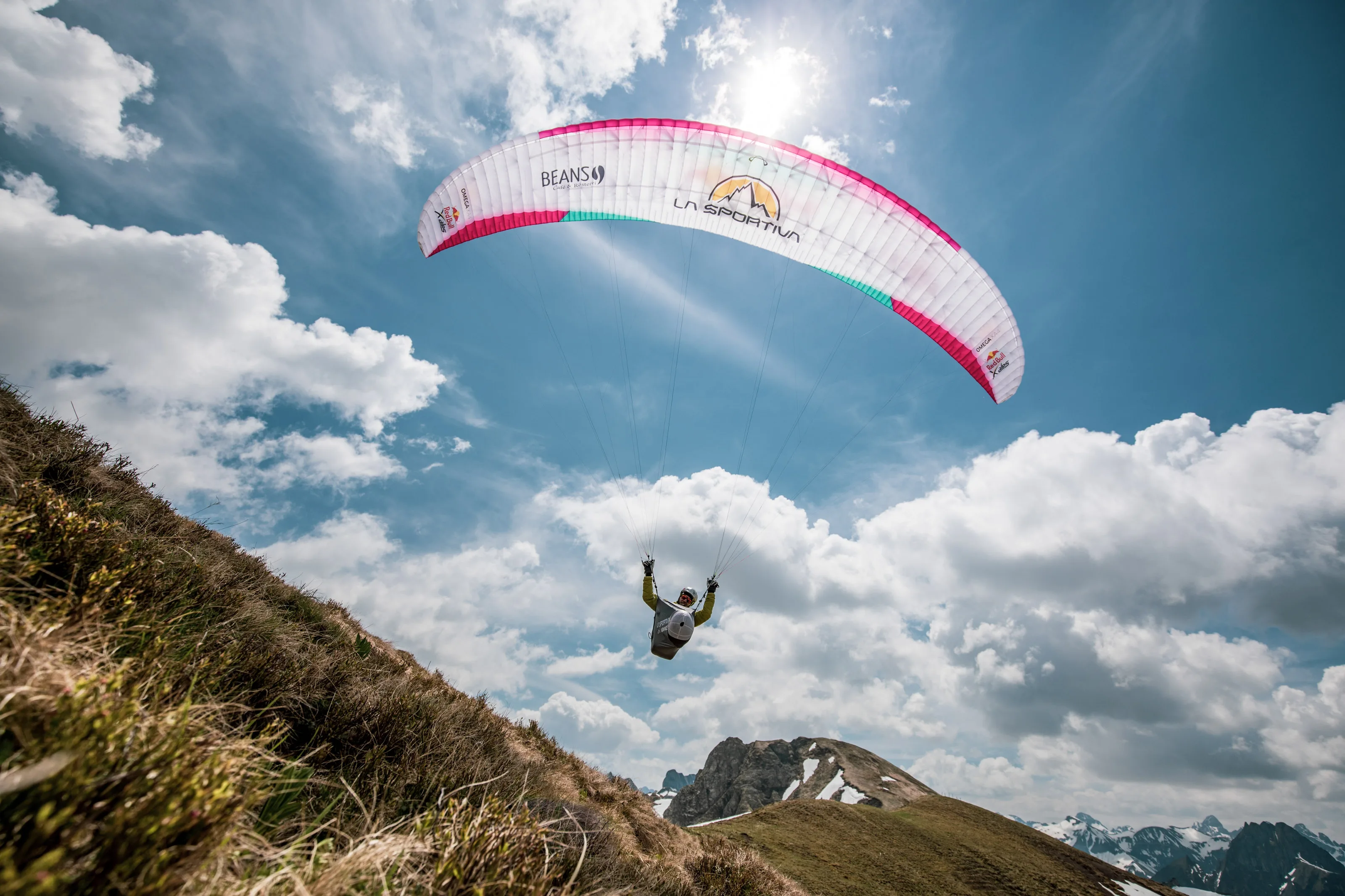 Ein pinkweißer Gleitschirm trägt Michael Lacher knapp über den Berghang vor weitem Himmel.