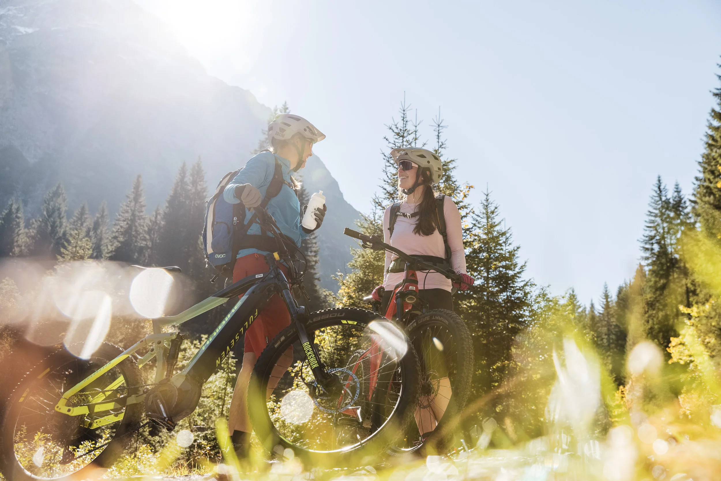 Silvi Vogel und Andrea Geiger stehen mit Mountainbikes im Gegenlicht der Berge