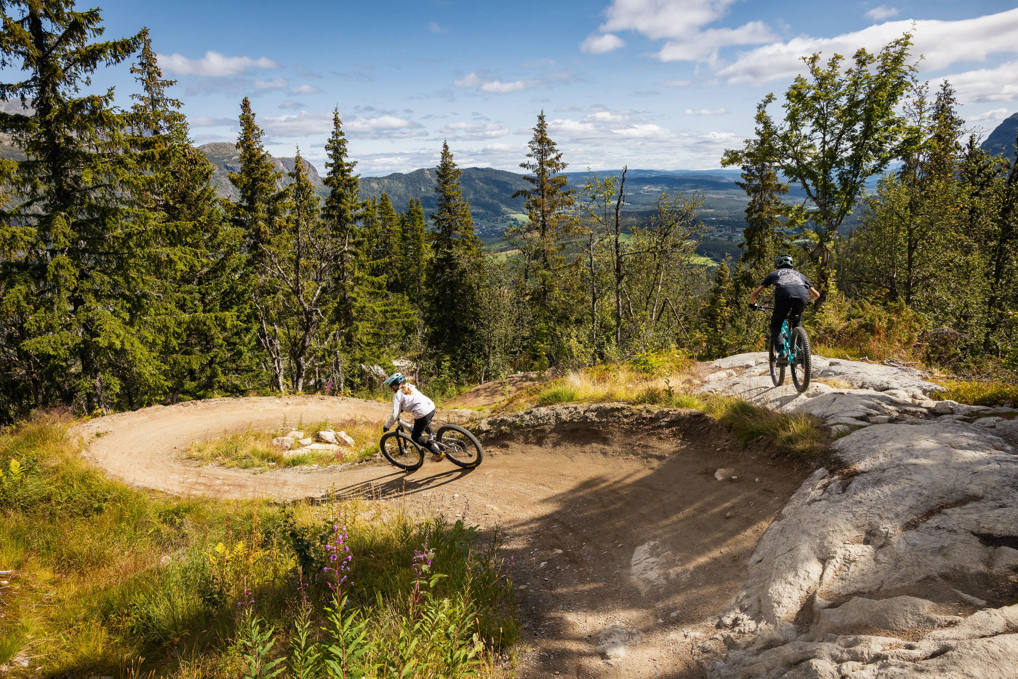 Zwei Mountainbiker folgen einander durch einen Waldanlieger mit felsiger Ausfahrt