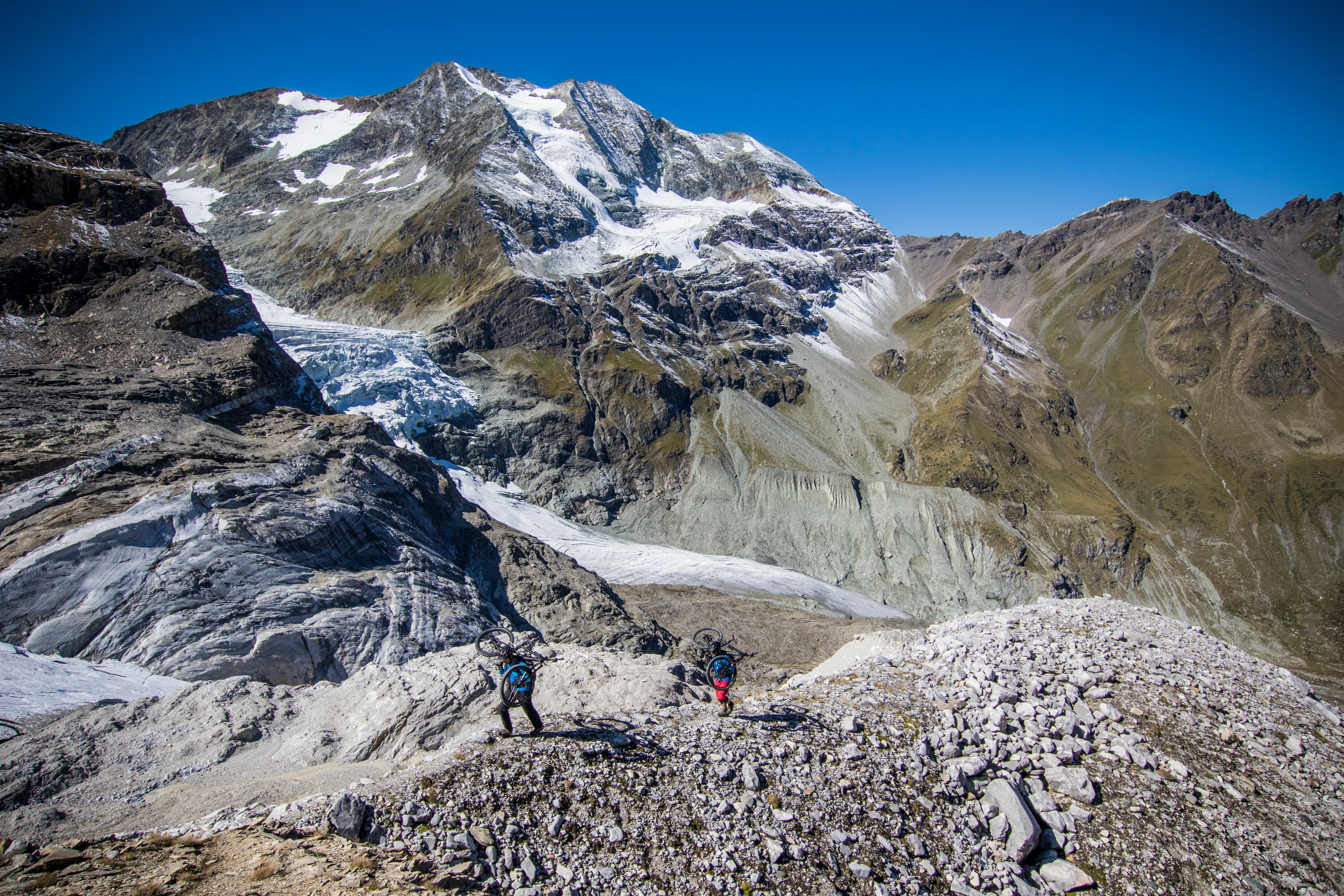 Mountainbiker bewegen sich mit ihren Rädern durch eine weite Geröllflanke im hochalpinen Wallis.