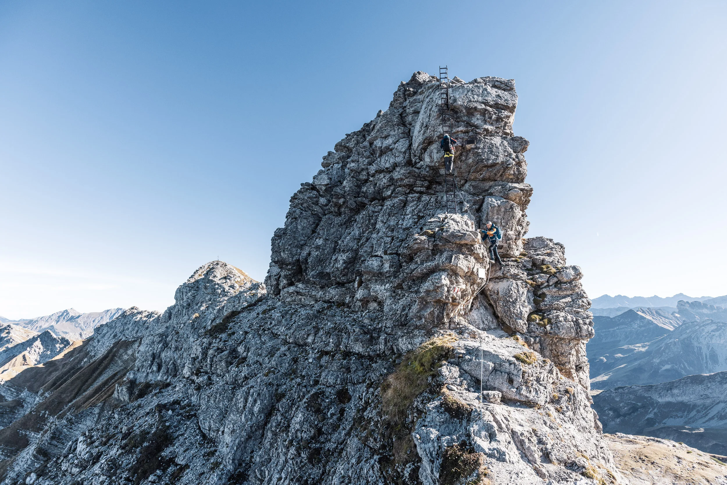 Zwei Bergsteiger klettern über Metallleitern an einem markanten Felsturm.